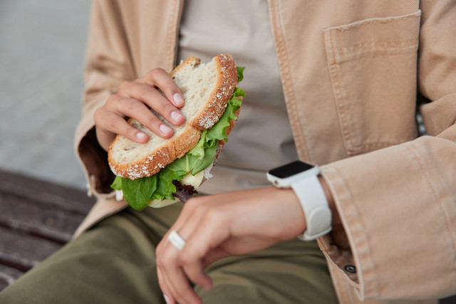 cropped-shot-faceless-peson-checks-time-digital-smartwatch-eats-delicious-sandwich-has-snack-rests-after-walking-sits-bench-chec